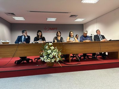 Foto de pessoas sentadas de forma enfileirada atras de uma mesa de madeira.